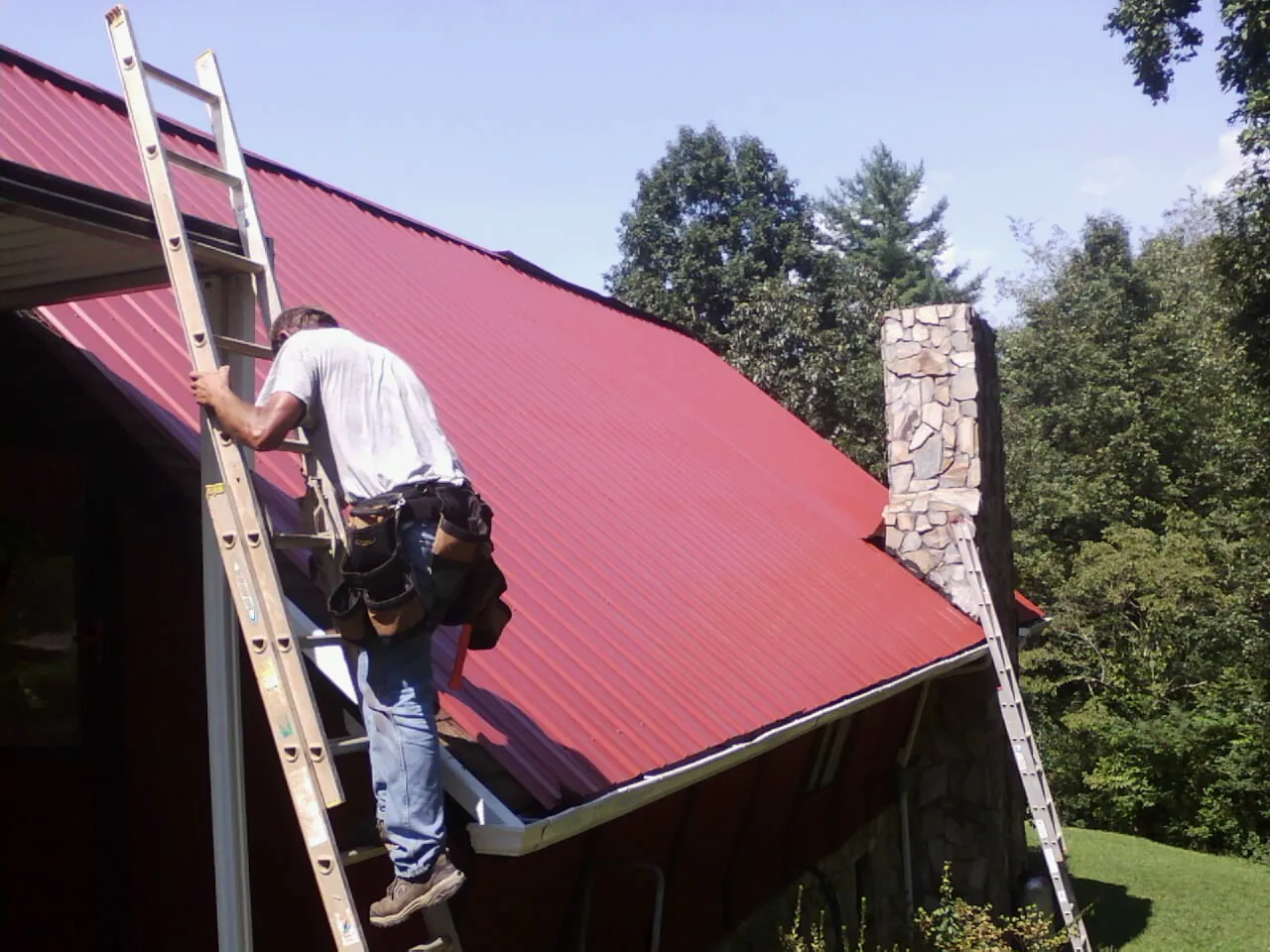Roofer on a ladder beside a red metal roof and stone chimney — Soffit & Fascia Repair by Roofing Brothers in Middle Point
